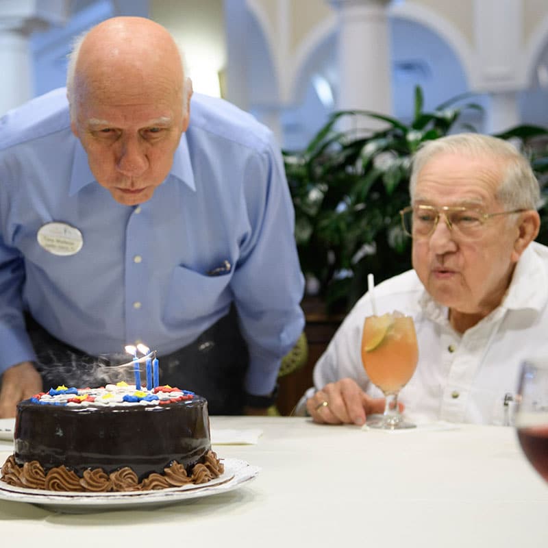 Two old men blowing out candles on a birthday cake.